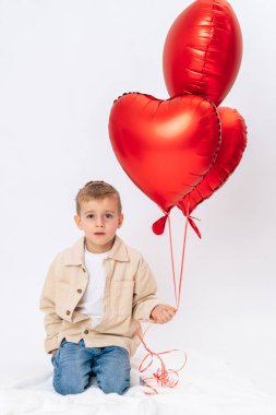Küçük çocuk kırmızı kalp şeklinde balonlarla beyaz arka planda diz çöküyor. Sevgililer Günü kutlaması, aşk sembolü, çocukluk sevinci, stüdyo resmi.