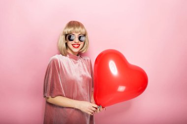 Seksi sarışın, kalp şeklinde sarılıyor kırmızı bir top ve gülümsüyor. Pembe bir arka planda.