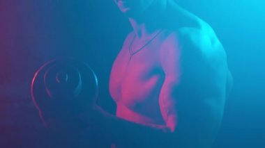 Stüdyodaki genç kaslı adam yeni ışıklar ve dumanla. Bir adam elinde dambıllarla kaslarını titretir..