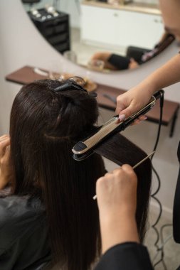 Professional hairstylist using a flat iron to straighten long dark hair in a beauty salon.