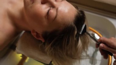 Bir kuaför saçını yıkarken genç bir kadın gözleri kapalı dinleniyor. Bir güzellik salonunda profesyonel saç bakımı ve kafa masajı.