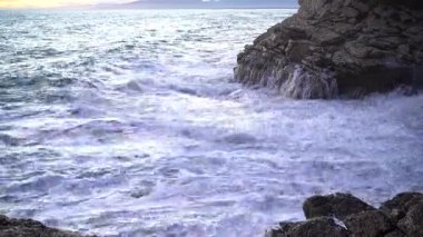 Engebeli bir uçurumun kenarına çarpıcı deniz dalgaları çarpıyor. Güzel ve sakin bir gün batımı arkaplanı olan manzaralı bir kıyı manzarası.