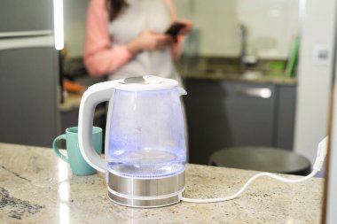 Mutfak tezgahındaki elektrikli su ısıtıcısı mavi ışıkla kaynayan su, arka planda akıllı telefon kullanan biri.