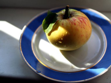 Elmanın sapında su damlaları ve yeşil yapraklar var. Pencere kenarında bir tabakta duruyor.