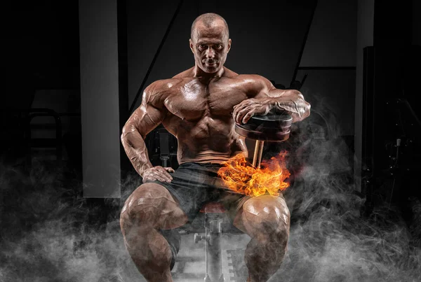 Muscular man sits on a bench with dumbbells burning. Bodybuilding and powerlifting concept. Mixed media