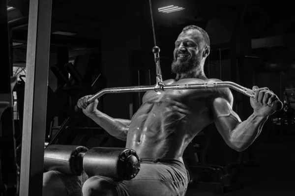 Professional athlete performs an exercise in the gym. Pulls the bar to his chest. Exercise for pumping the back. Mixed media