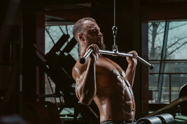 Professional athlete performs an exercise in the gym. Pulls the bar to his chest. Exercise for pumping the back. Mixed media