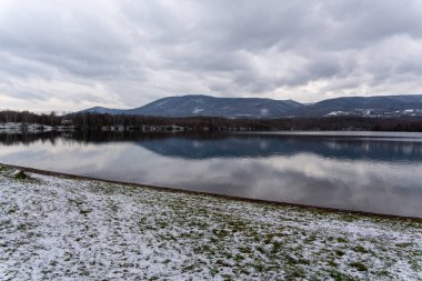Sakin gölü, dağları ve bulutlu gökyüzünü yansıtan huzurlu kış manzarası. Ön planda karlı bir kıyı şeridi, su boyunca çıplak ağaçlar, ve yumuşak, sessiz kış renkleri sakin, doğal bir hava yaratır.