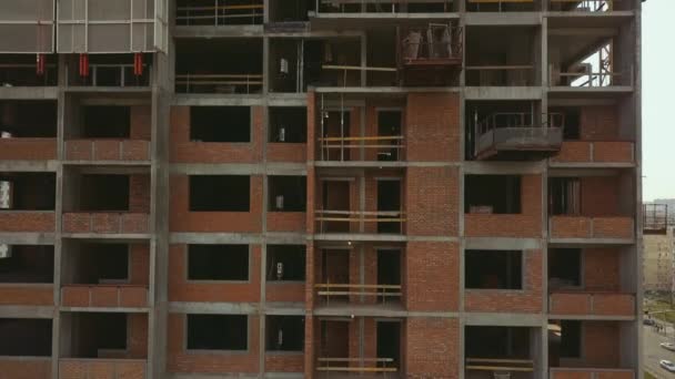 Levantando la cámara a lo largo de un edificio en construcción con espacios vacíos y trabajadores en el techo con la luz contrastante del sol — Vídeo de stock