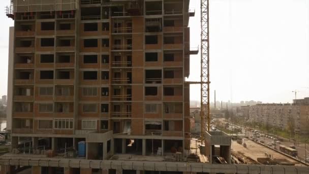 Levant la caméra le long d'un bâtiment en construction avec des travées de plancher vides et des travailleurs sur le toit avec la lumière contrastée du soleil — Video