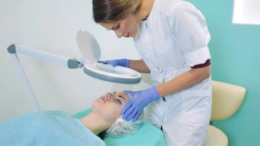 Güzellik uzmanı muayenehanede bir kızın derisini büyüteçle inceliyor. Kadın bir kozmetikçinin işi..