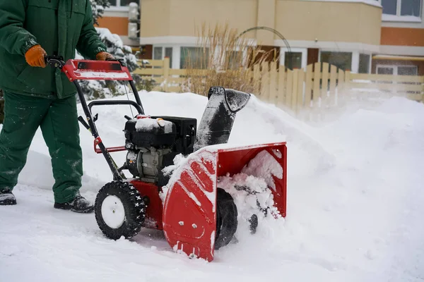 Pracownik w zielonym kombinezonie usuwa śnieg czerwoną dmuchawą na tle świerku, zaspy śnieżnej i domu. Obrazy Stockowe bez tantiem