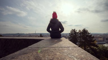 Kışlık ceketli kadın Letna parkında duvarda oturuyor ve Prag 'ın manzarasına bakıyor.