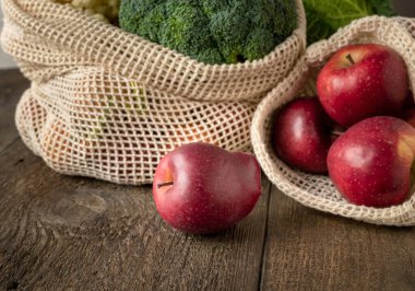 Tahta bir masada organik pamuktan yapılmış ekolojik yeniden kullanılabilir ağ torbalarında taze elmalar ve brokoli.