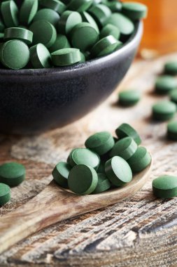 Bir kaşıkta ve siyah bir kasede yeşil spirulina alg tabletleri.