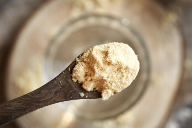 Tahta bir kaşıkta beyaz protein tozu, bir bardak suyun üstünde, üst manzara.