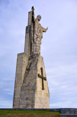 Düşük açılı, anıtsal taş İsa heykeli. Kolları açık ve mavi gökyüzünün altındaki yıpranmış kaidede çapraz.