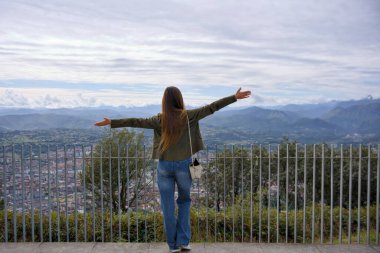 Bulutlu bir günde, şehre ve sıradağlara tepeden bakan geniş kolları olan bir kadının dikiz görüntüsü. 