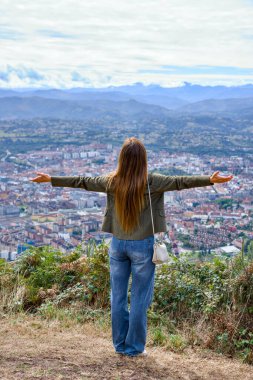 Kolları ardına kadar açık, şehir vadisi ve dağların panoramik manzarasının tadını çıkaran kadın yolcunun arkası.