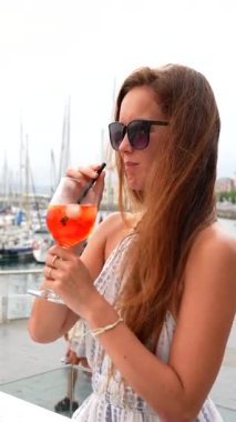 Güneş gözlüklü genç bir kadın arka planda yat ve liman olan Gijon İspanya 'da açık yat kafesinde saman yoluyla portakal kokteyli içiyor.