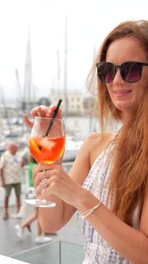 Güneş gözlüklü genç kadın elinde turuncu kokteyl bardağıyla marinada rahatlatıcı yaz anının tadını çıkarıyor. Arka planda yatları olan Gijon İspanya.