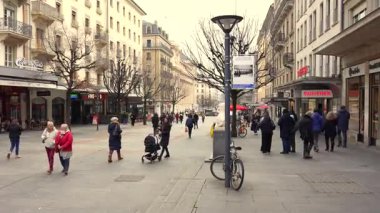 17 Şubat 2025 'te Cenevre' de, ağaçsız kafeler ve perakendeciler arasında dolaşan insanlarla dolu bir cadde.