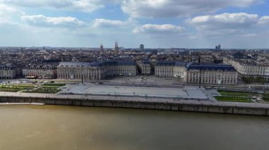 Bordeaux Place de la Bourse 'un insansız hava aracı videosu, tarihi taş binalar, yayalarla dolu geniş bir meydan ve yumuşak bahar güneşi altında çamurlu Garonne nehri.
