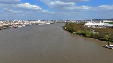 Bulutlu bir bahar gününde Bordeaux 'daki yeşil nehir kenarı parkı ve modern binalar boyunca yanaşan gemilerin insansız hava aracı videosu.