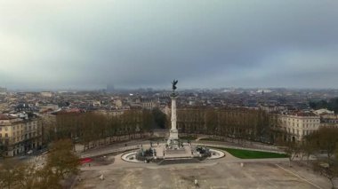 Esplanade des Quinconces 'daki ağaçların ve tarihi binaların üzerindeki melek heykeli ve bulutlu gökyüzü altındaki Anıt aux Girondins' in insansız hava aracı görüntüleri.