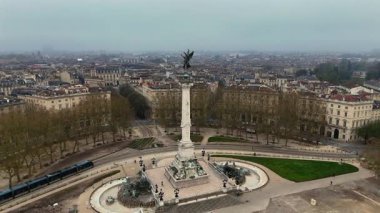 Monument aux Girondins 'in insansız hava aracı çekimi ve içinden geçen tramvay, çıplak ağaçlar ve yumuşak sabah sisi içinde tarihi şehir silueti olan çeşme.