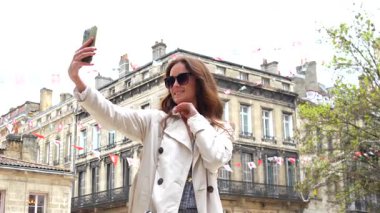 Güneş gözlüklü kadın Bordeaux Meydanı 'nda taş binalar ve bayraklarla selfie çekiyor.