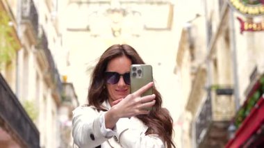 Güneş gözlüklü kadın Bordeaux caddesinde balkonlu ve tabelalı selfie çekiyor.
