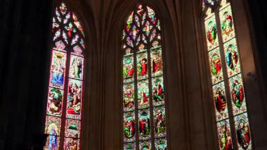 Gotik katedral iç mimarisi, sıcak ışık altında incilsel sahneleri gösteren vitray pencereli.