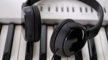 Dijital piyano klavyesinde dinlenen siyah kulaklıklar modern müzik yaratılışını, ses kaydını ve çevrimiçi öğrenmeyi sembolize ediyor. Müzik eğitimi, dijital piyano uygulaması, ses üretimi, podcast ve rel kavramlarını resmetmek için ideal