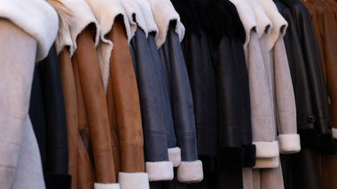 Bir sıra şık koyun derisi ve çeşitli renklerde deri paltolar askıda asılı duruyor. Görüntü, moda, perakende satış ve mevsimlik giysi kavramları için ideal doğal malzemelerden yapılmış ılık kış kıyafetlerini sergiliyor..