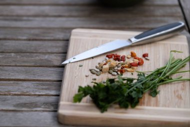 Taze maydanozun yakın plan görüntüsü ve doğrama sebzeler. Dışarıdaki ahşap bir kesme tahtasının üzerinde paslanmaz çelik bir mutfak bıçağıyla. Sağlıklı yemek yapma, doğal malzemeler ve dışarıda yemek hazırlama kavramı. Tarif siteleri, yemek blogları ve beslenme için ideal