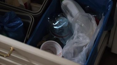 Kadınların mavi bir çantayı plastik atıklarla dolu bir çantayla geri dönüşüm çekmecesine bağladığı yakın çekim videosu. Arka planda kağıt ve karışık atık için diğer kutular görülebilir. Ev geri dönüşümü kavramı