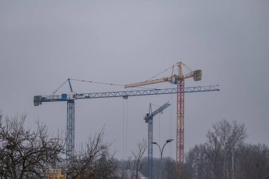 Kış kentlerinin üzerinde çıplak ağaçlar ve bulutlu gökyüzü ile birden fazla kule vinci yükseliyor. Sahne aktif inşaat, gayrimenkul geliştirme ve şehir genişlemesini temsil ediyor. Temalar için uygun