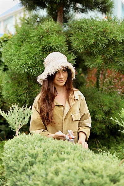 Дівчина садівник в робочому одязі і солом'яному капелюсі вирізає садові ножиці вічнозелені — стокове фото