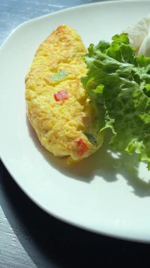 Sarı omlet ve yeşil yapraklı sebzeli bir tabak yemek. Tabak beyaz ve yemek düzgünce düzenlenmiş. Yüksek kalite 4k görüntü