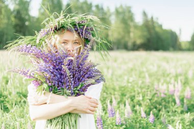 Elinde bir buket kır çiçeği tutan hippi bir kız. Kız yüzünü lupin buketinin arkasına sakladı. Kız çiçek tarlasında büyük bir buket mor lupin tutuyor. Doğa konsepti. Burada.