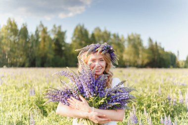 Elinde bir buket kır çiçeği tutan hippi bir kız. Kız yüzünü lupin buketinin arkasına sakladı. Kız çiçek tarlasında büyük bir buket mor lupin tutuyor. Doğa konsepti. Burada.