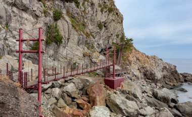 Güney Kore 'nin Ulleungdo Adası' ndaki kayalık kıyı şeritleri ve mavi okyanus suları boyunca kırmızı kahverengi asma köprü yolu.