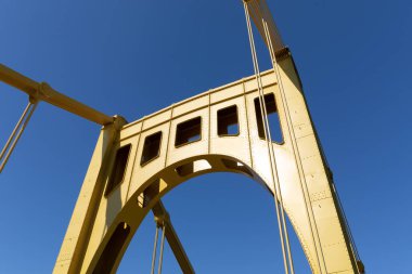 Kendi kendine sabitlenmiş asma köprünün sağ tarafı açık sarıya boyalı derin mavi bir gökyüzüne, yatay bir görünüme