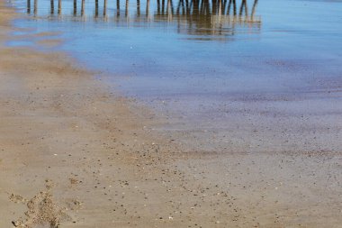 Mavi gökyüzü kumlu bir plajın kenarındaki suya yansıyor, dar iskele kazıkları, yatay görünüm