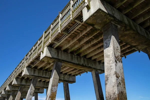 海の桟橋の下からの眺め 木製の桟橋デッキと手すりを支えるコンクリート製の杭 深い青い空 水平方向の側面にシルエット — ストック写真