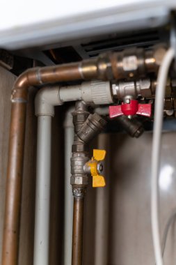 Kazanın altındaki tesisat bağlantılarının yakından görüntüsü, metal boruları, vanaları ve tesisatları gösteriyor. Resim ev bakımını, ısıtma sistemlerini ve temel araç altyapısını vurguluyor