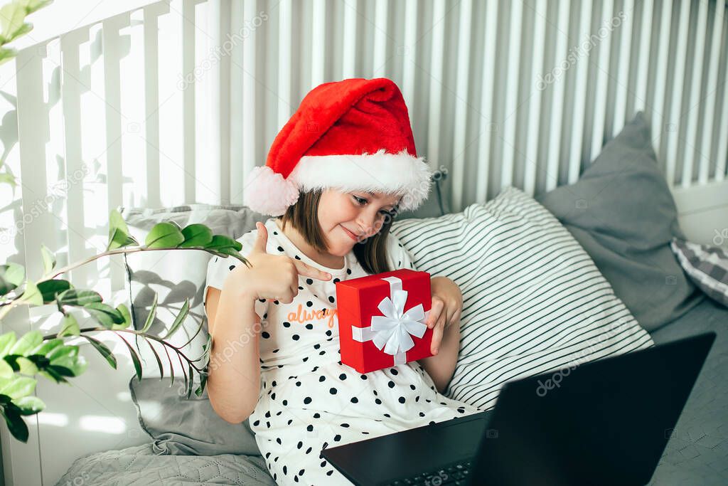 Girl in hat of Santa Claus holding gift box and communicates with friends via video chat. COVID, social distancing concept