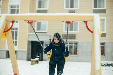 Kış kıyafetli küçük çocuk kar yağarken sallanıyor. Çocuk dışarıda oyun parkında oynuyor. Kış tatili ve mevsim konsepti