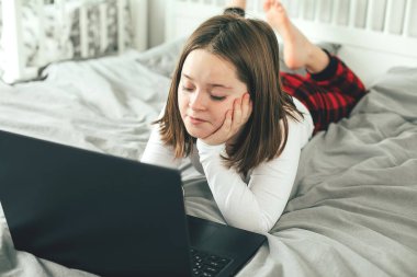 Genç kız yatakta oturuyor ve dizüstü bilgisayar kullanıyor. Çevrimiçi baskı kavramı
